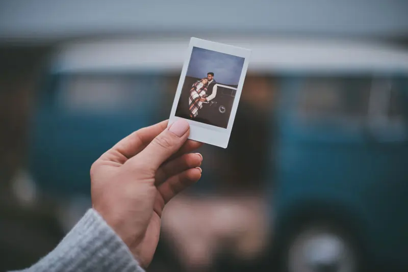Eine Hand hält ein altes Polaroid-Foto eines Paares vor einem verschwommenen Hintergrund, das Erinnerungen an gemeinsame Zeiten zeigt.